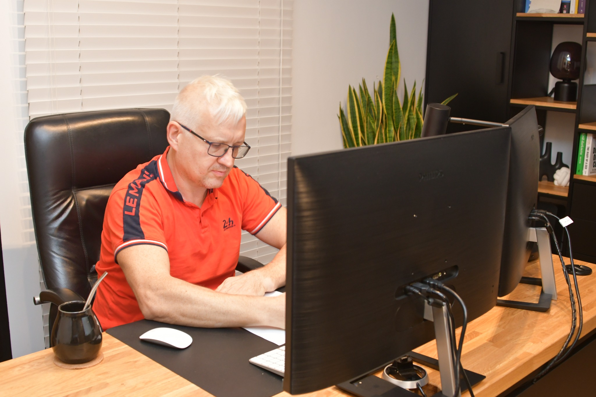 Mężczyzna w okularach siedzi przy biurku z dwoma monitorami, klawiaturą i myszką, obok stoi naczynie z yerba mate, w tle roślina doniczkowa i regał.