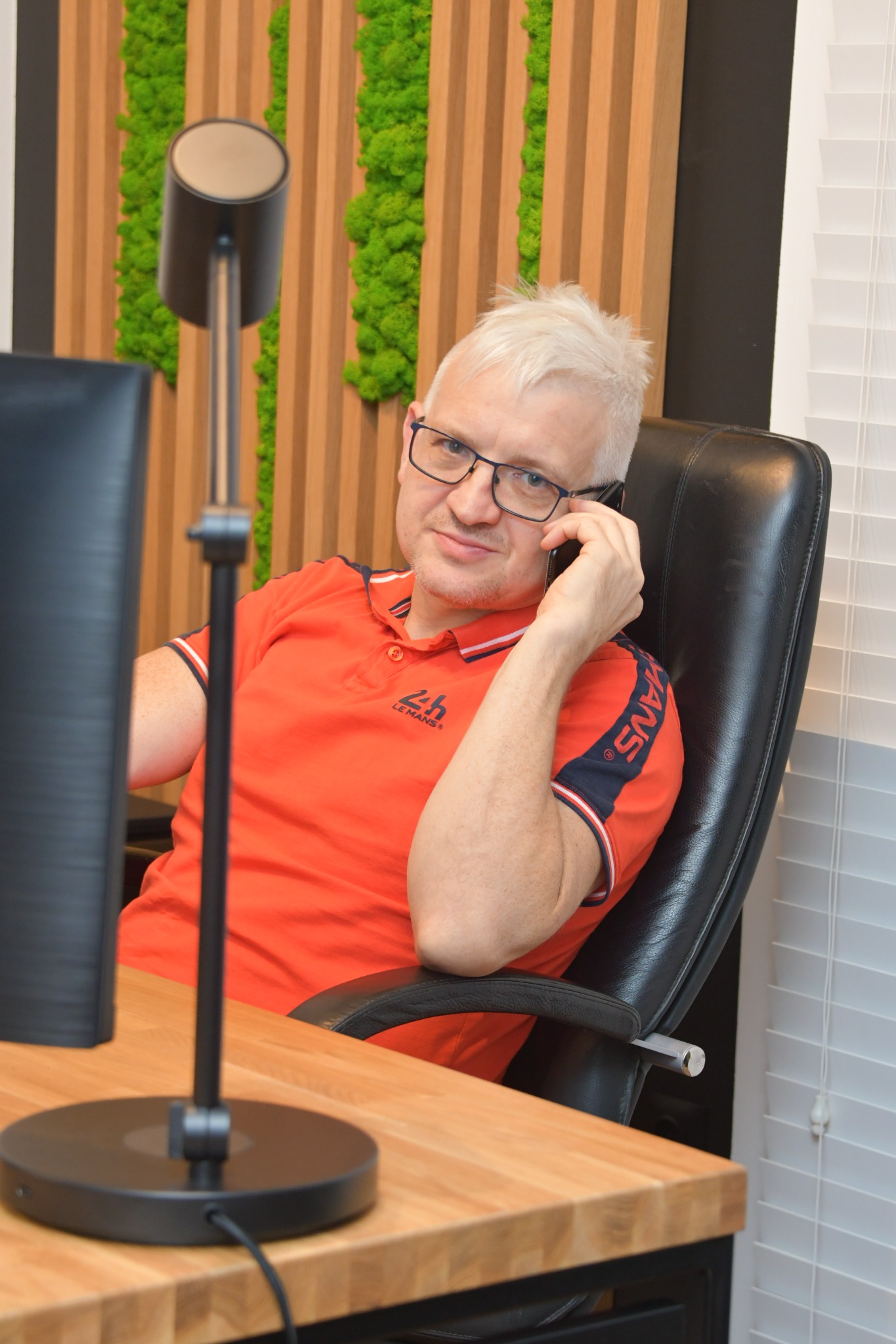 Mężczyzna w okularach i pomarańczowej koszulce siedzi przy biurku, rozmawia przez telefon, obok monitor i lampka biurkowa; w tle drewniana ściana z panelami z mchu.