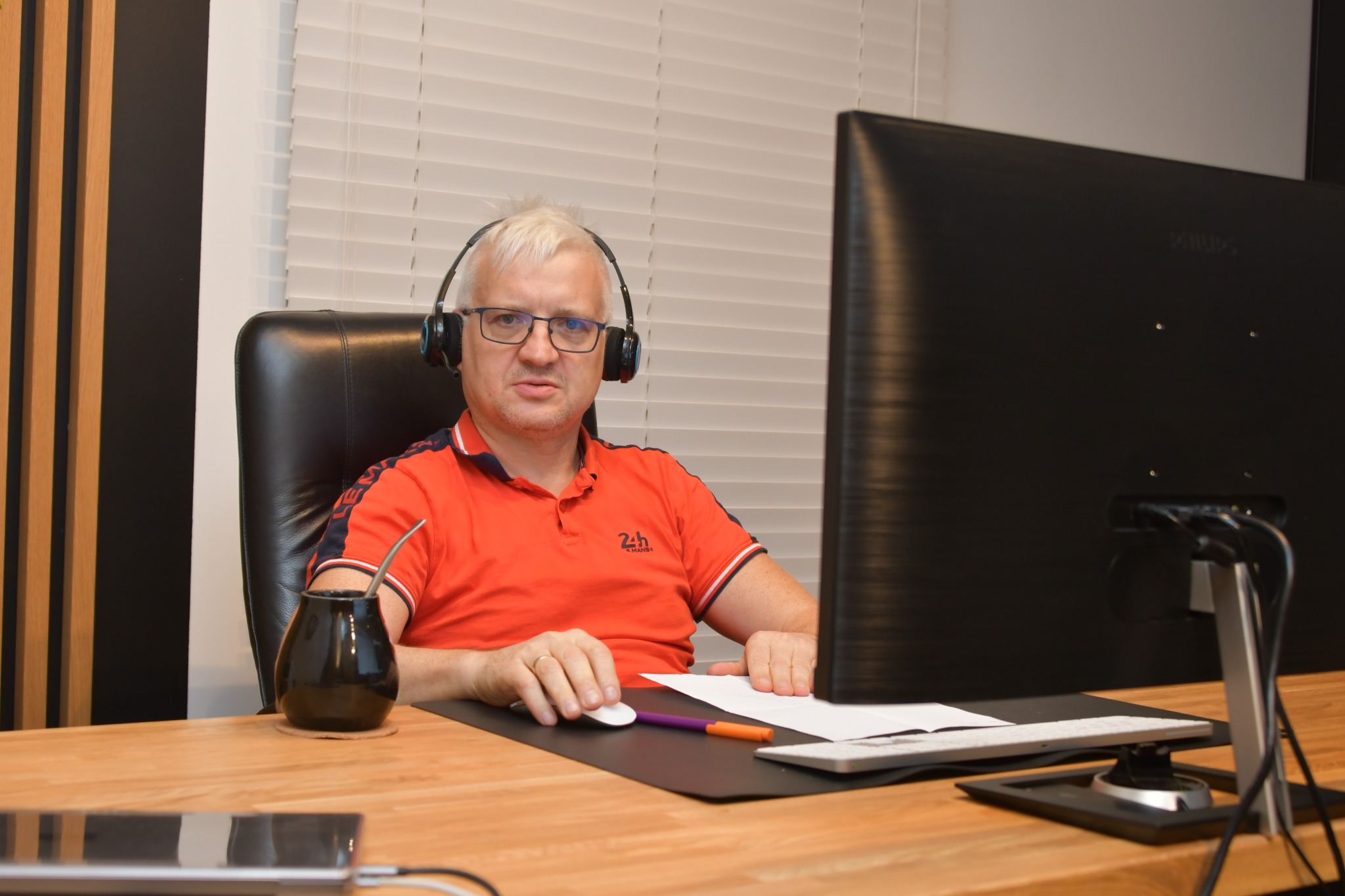 Mężczyzna w okularach i słuchawkach siedzi przy biurku z dużym monitorem, używając myszki komputerowej, obok yerba mate i dokumenty.