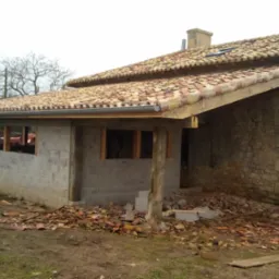 Budynek w trakcie remontu dachu z widocznymi starymi dachówkami na ziemi, ściany częściowo z bloków betonowych, a częściowo z kamienia.