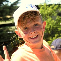 Agencja Turystyczno-Artystyczna Przygoda Anna Gołębiowska-Rusak - Uśmiechnięty chłopiec z trawą we włosach i na twarzy, ubrany w czapkę i pomarańczową koszulkę, pokazuje znak pokoju na tle zieleni.