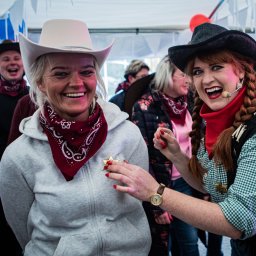Agencja Turystyczno-Artystyczna Przygoda Anna Gołębiowska-Rusak - Uśmiechnięte kobiety w strojach kowbojskich podczas imprezy tematycznej, jedna przypina drugiej gwiazdę szeryfa, w tle widoczni inni uczestnicy zabawy i balony.