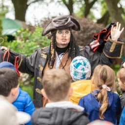 Osoba przebrana za pirata, z charakterystycznym makijażem i kostiumem, prezentuje strój dzieciom podczas wydarzenia plenerowego.