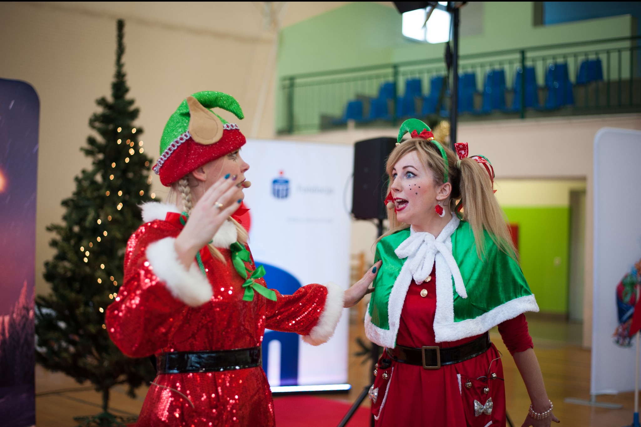 Dwie kobiety przebrane za elfy w błyszczących kostiumach, z mikrofonami nagłownymi, prowadzą interakcję na tle choinki i sali gimnastycznej.