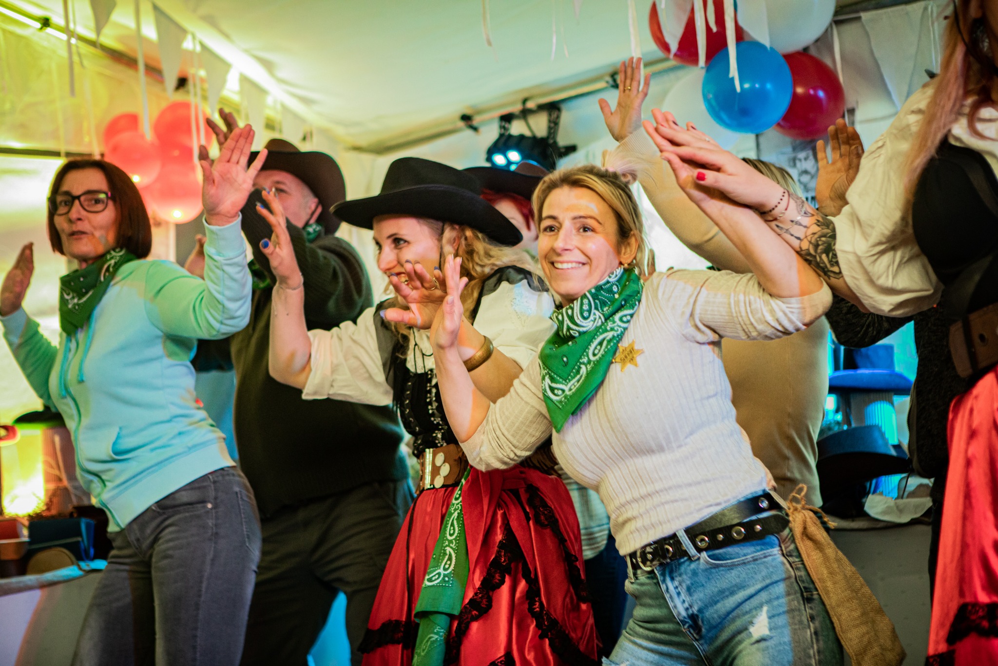 Grupa uśmiechniętych osób w strojach kowbojskich tańczy w oświetlonym namiocie z balonami, widoczne detale kostiumów, takie jak bandany i gwiazdy szeryfa.