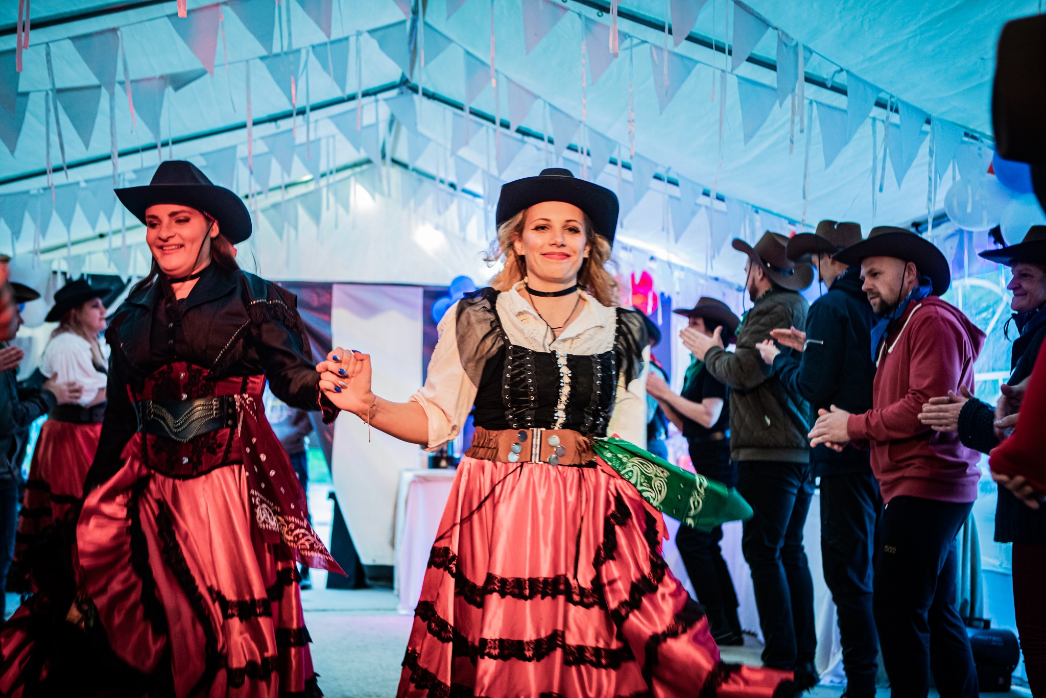 Dwie kobiety w kowbojskich strojach taneczne na scenie podczas wydarzenia, w tle publiczność w kowbojskich kapeluszach klaszcze, dekoracje z białych chorągiewek pod sufitem.