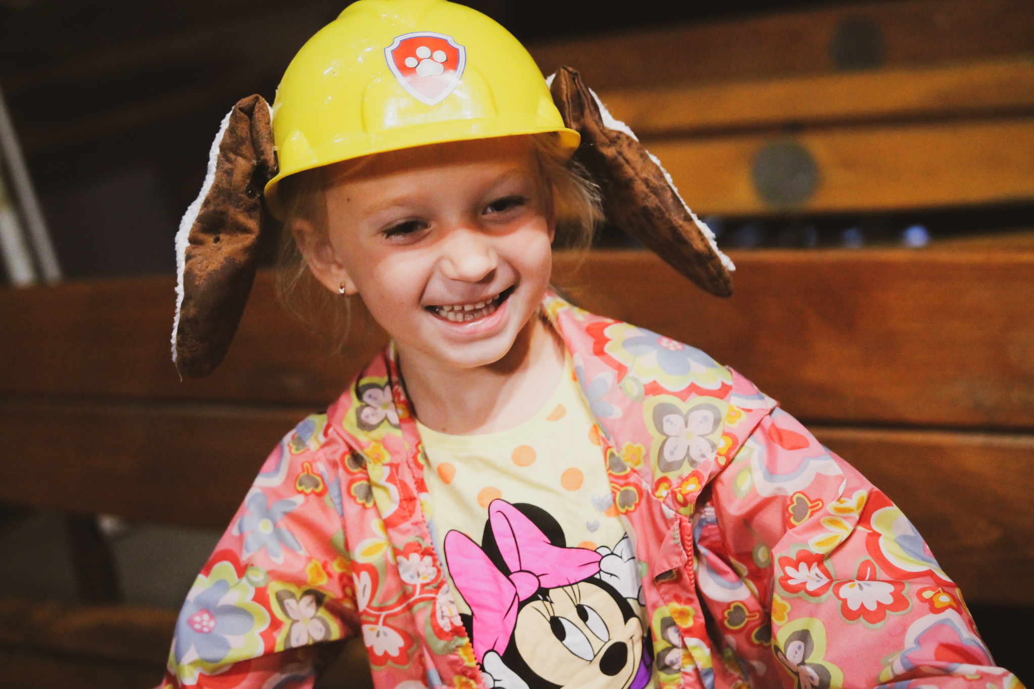 Uśmiechnięta dziewczynka w żółtym kasku z uszami psa i logiem łapy, ubrana w bluzkę z Minie i kolorową kurtkę na tle drewnianej ściany.
