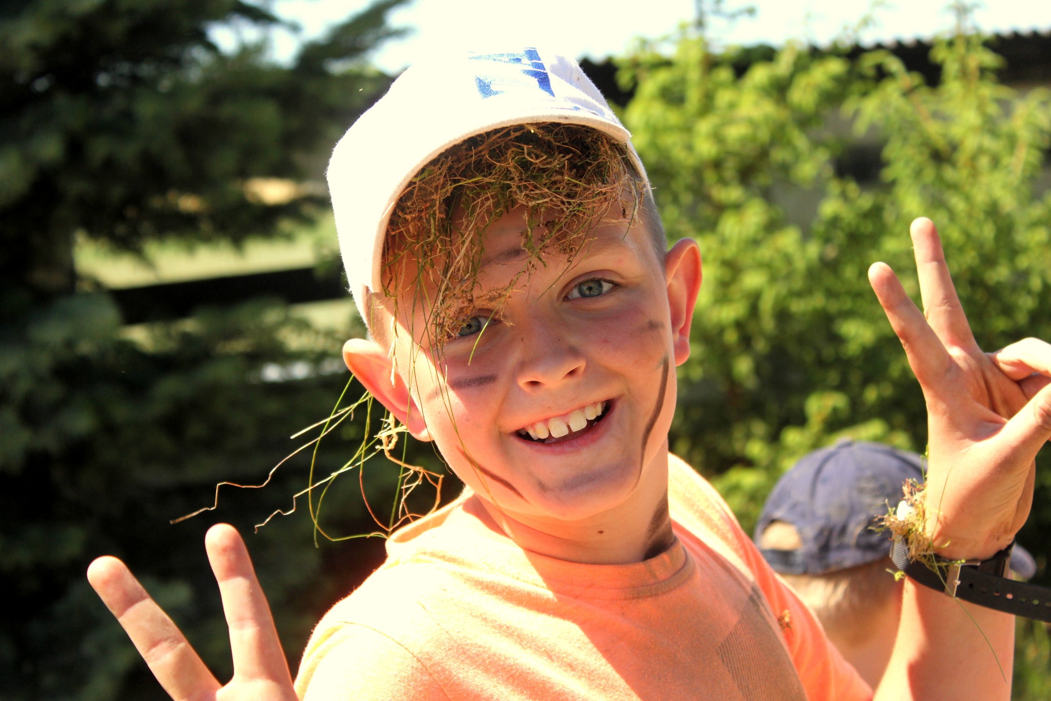 Uśmiechnięty chłopiec z trawą we włosach i na twarzy, ubrany w czapkę i pomarańczową koszulkę, pokazuje znak pokoju na tle zieleni.