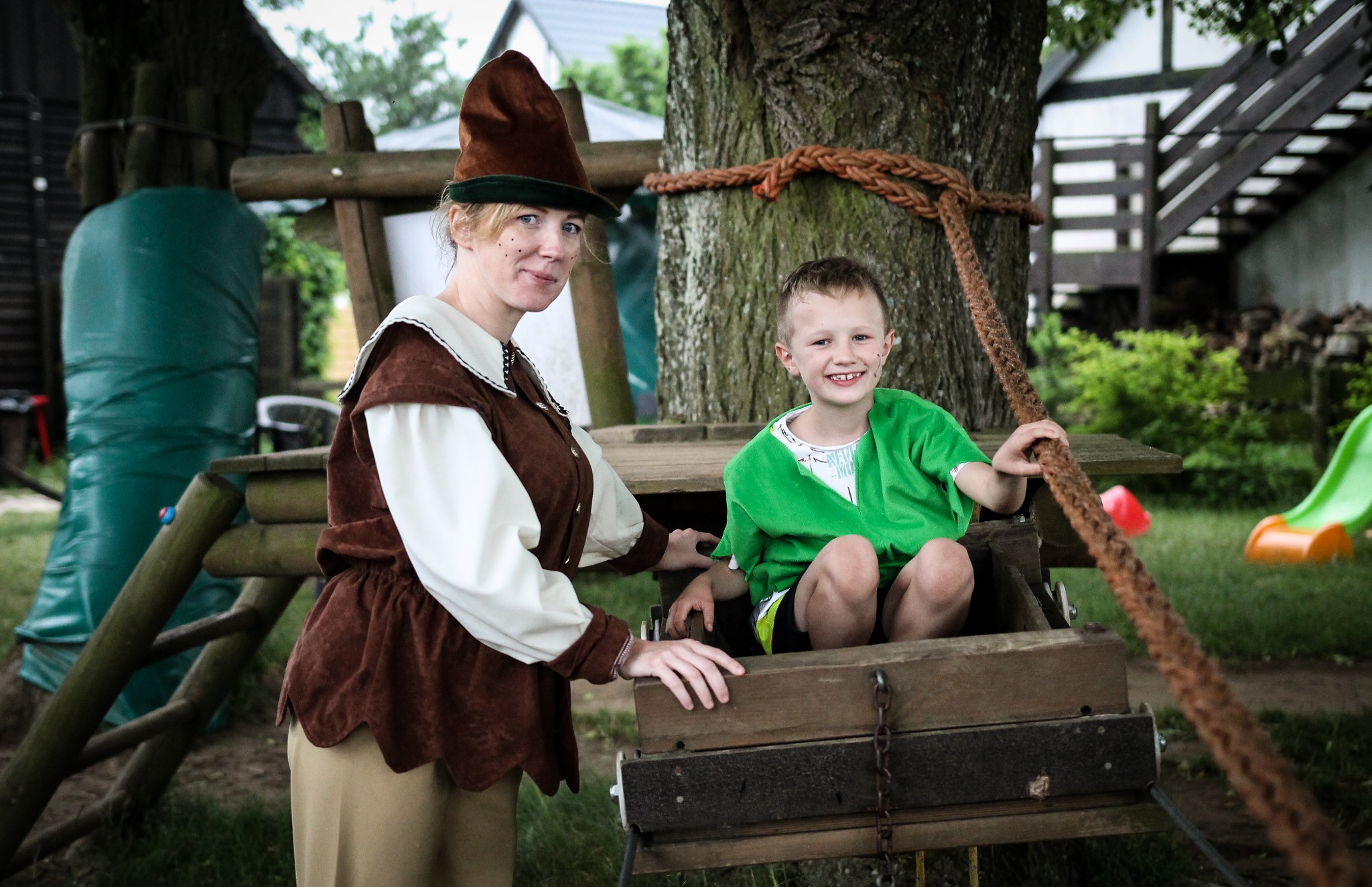 Kobieta w kostiumie i uśmiechnięty chłopiec w zielonej koszuli siedzą w drewnianej skrzyni zawieszonej na linie przy drzewie, z elementami placu zabaw w tle.