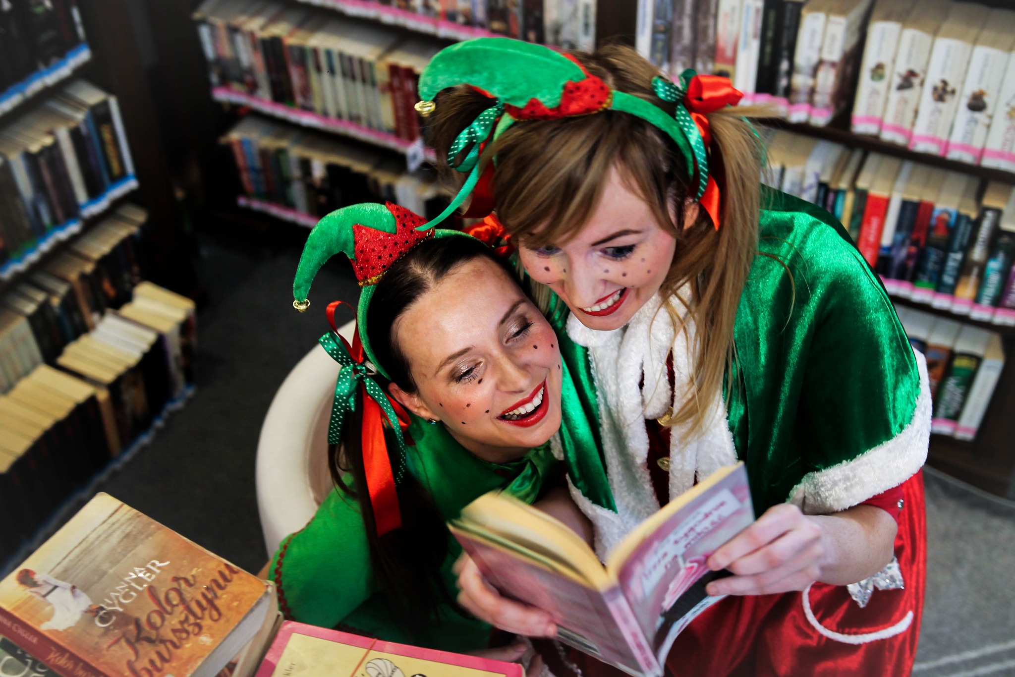Dwie uśmiechnięte kobiety przebrane za elfy czytają książkę w bibliotece, z charakterystycznymi elfimi czapkami i czerwonymi ustami.
