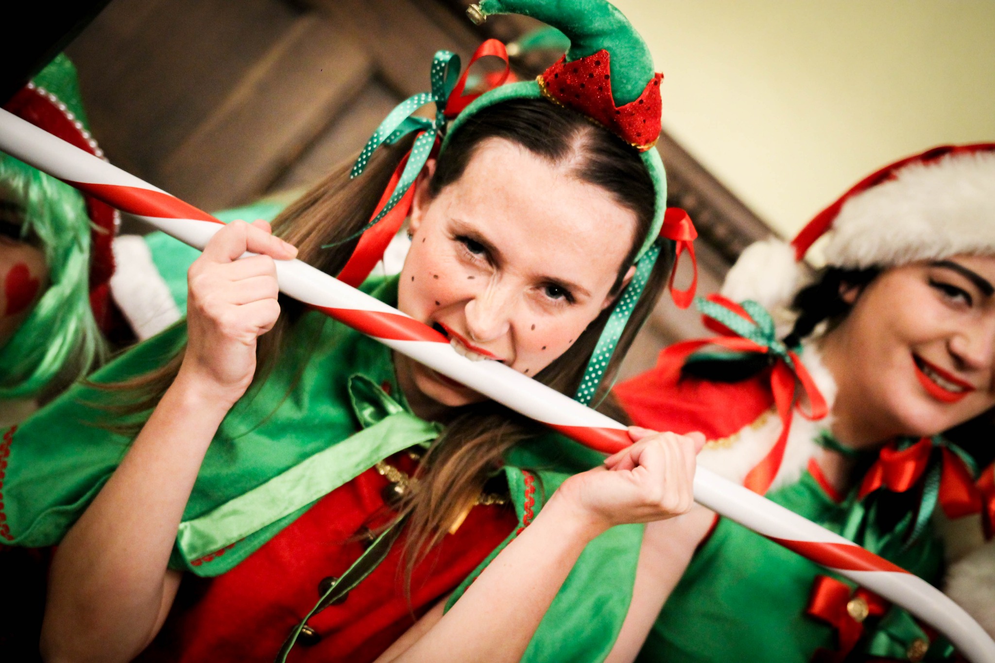 Kobieta w stroju elfa z zielonym kapeluszem i czerwonymi akcentami, gryząca biało-czerwoną laskę cukrową, z uśmiechem i pomalowanymi piegami, obok druga osoba w stroju Mikołaja.