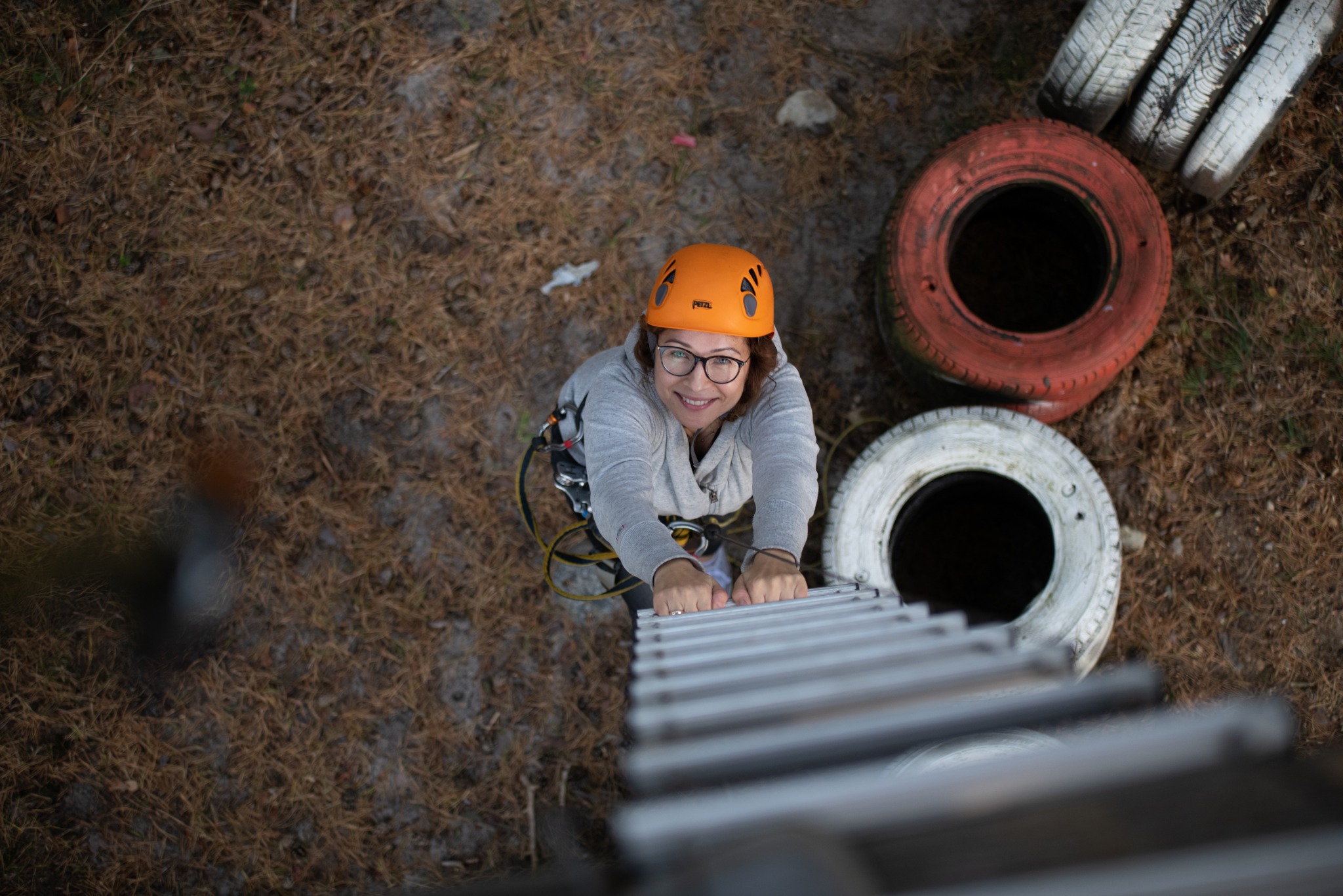 Kobieta w pomarańczowym kasku wspina się po drabince na tle leśnej ściółki i opon pomalowanych na biało i czerwono.