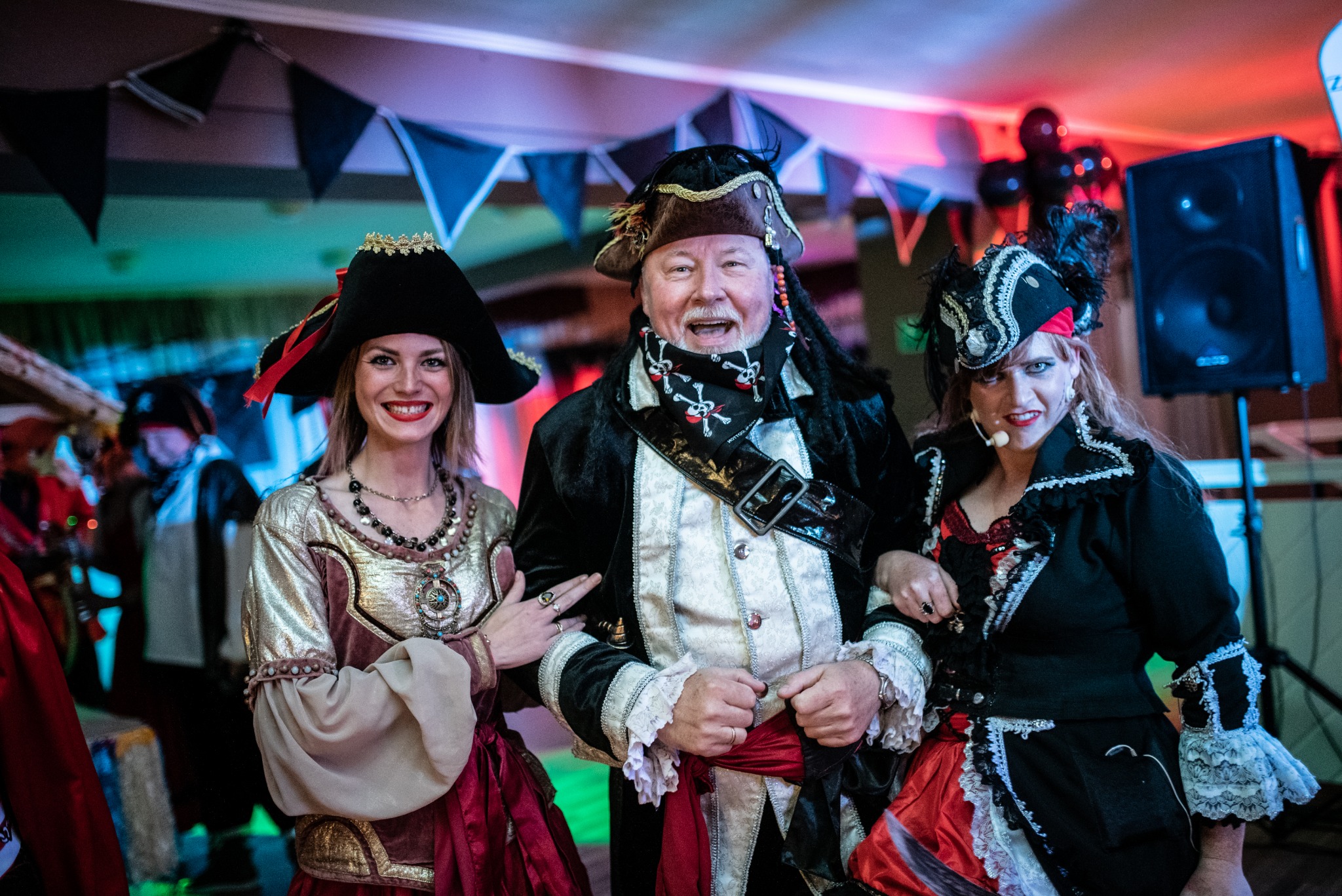 Trzy osoby przebrane w stroje pirackie podczas tematycznego przyjęcia, widoczne dekoracje i nagłośnienie.