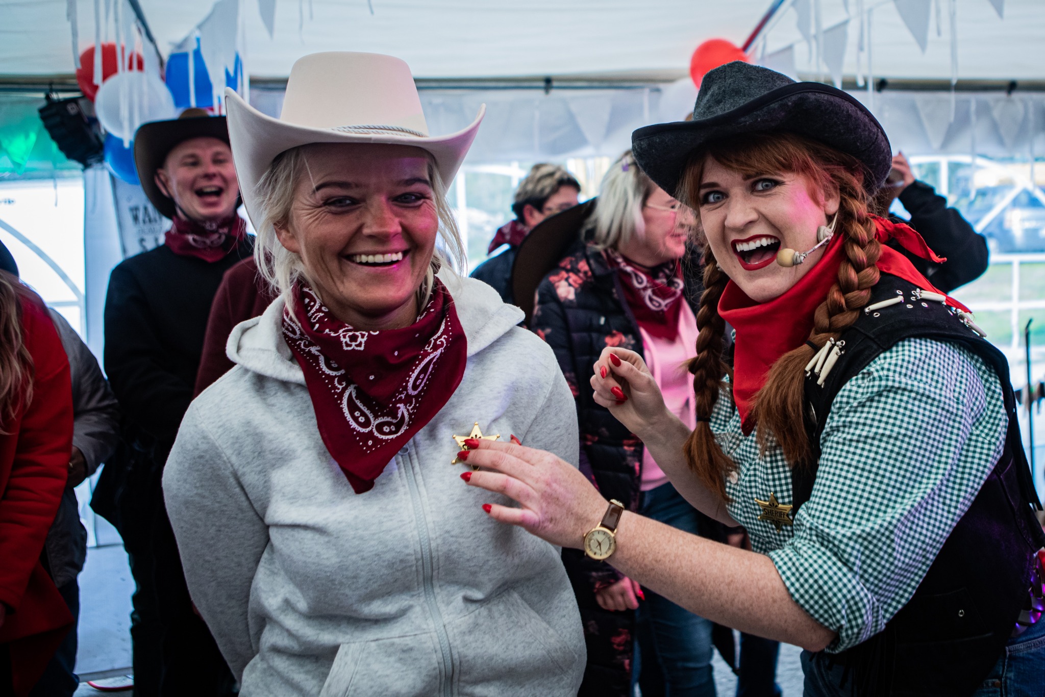 Uśmiechnięte kobiety w strojach kowbojskich podczas imprezy tematycznej, jedna przypina drugiej gwiazdę szeryfa, w tle widoczni inni uczestnicy zabawy i balony.