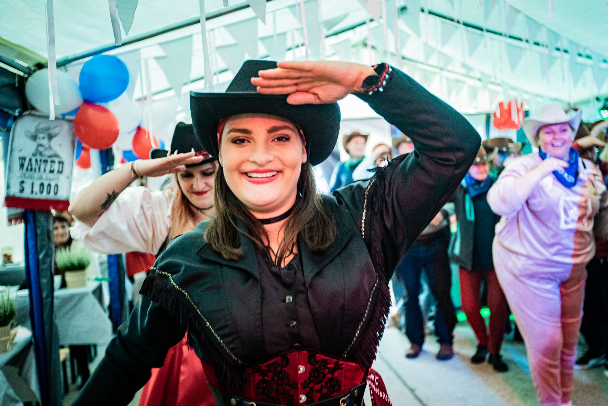 Uśmiechnięta kobieta w stroju kowbojskim i czarnym kapeluszu salutuje na tle tłumu osób również przebranych w stroje westernowe, dekoracje z balonów i girlandy tworzą imprezową atmosferę.