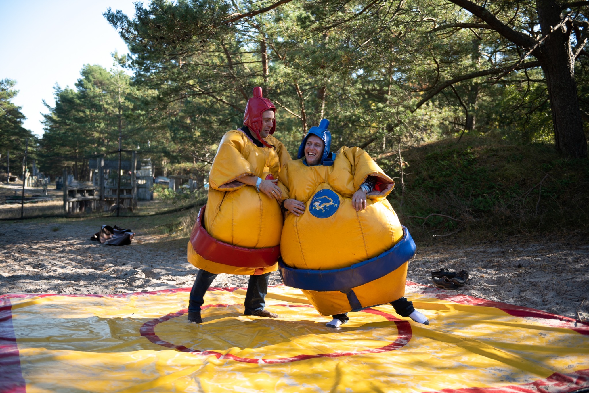 Dwie osoby w kostiumach sumo na dmuchanej macie, rozgrywka w plenerze, w tle widoczne drzewa i elementy konstrukcji.