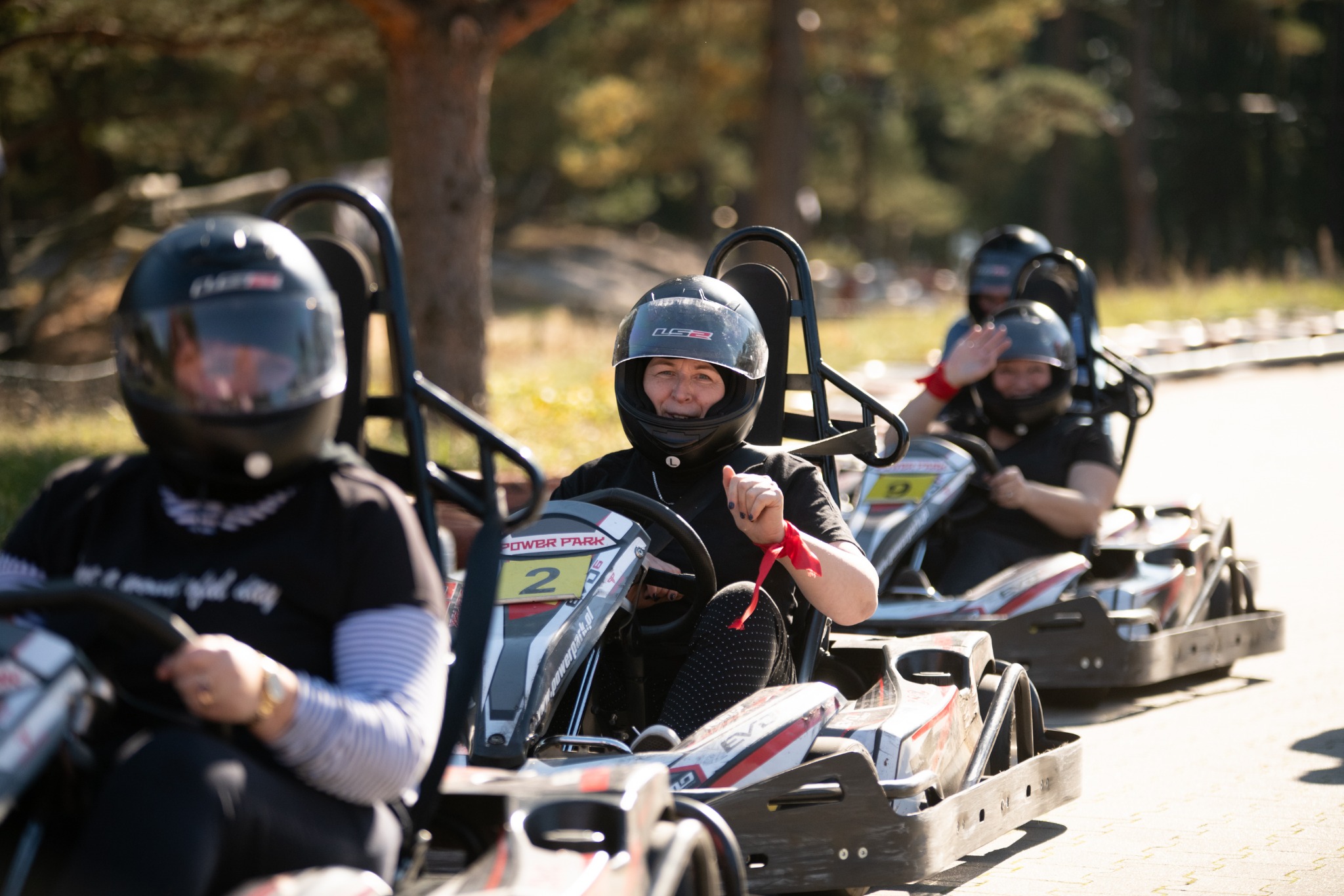 Uśmiechnięta kobieta w kasku siedzi w gokarcie oznaczonym numerem 2 na torze gokartowym, w tle widoczni inni uczestnicy zabawy.