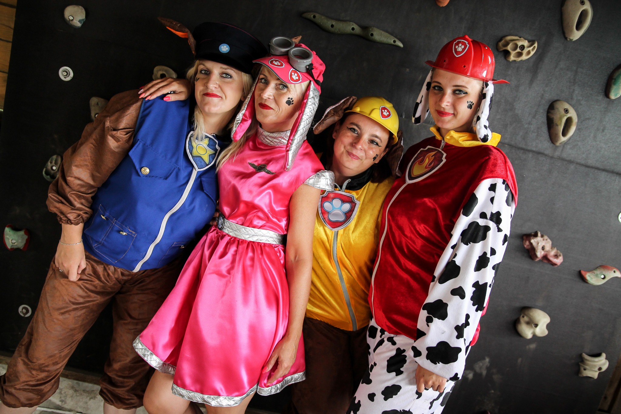Cztery kobiety w kostiumach inspirowanych Psim Patrolem, pozujące na tle ścianki wspinaczkowej z uchwytami.