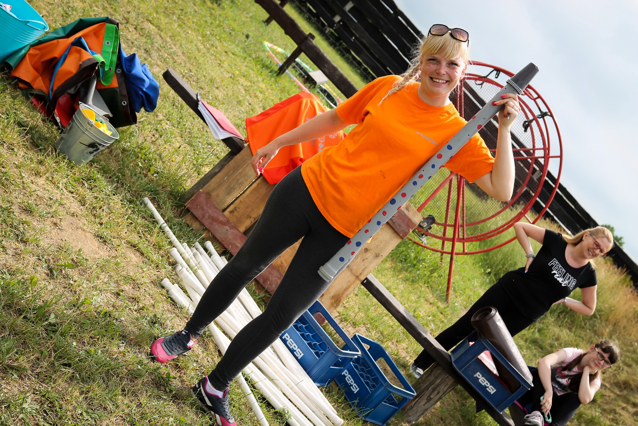 Uśmiechnięta blondynka w pomarańczowej koszulce trzyma szarą rurę z kolorowymi kropkami na tle trawy i rekwizytów do zabaw na świeżym powietrzu, w tle widoczna konstrukcja z czerwonych prętów.