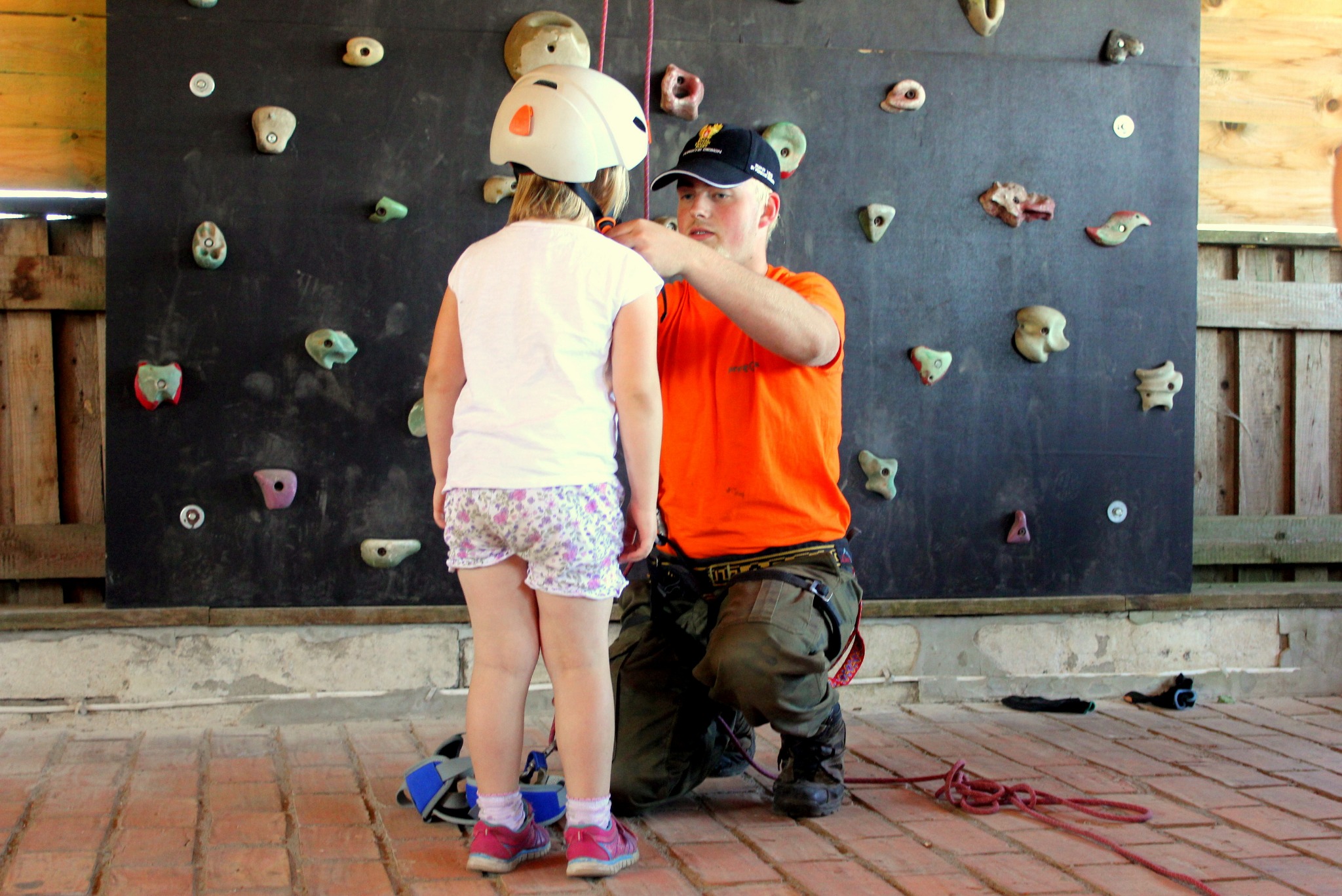Instruktor dopasowuje kask dziewczynce przed ścianką wspinaczkową z różnorodnymi uchwytami.