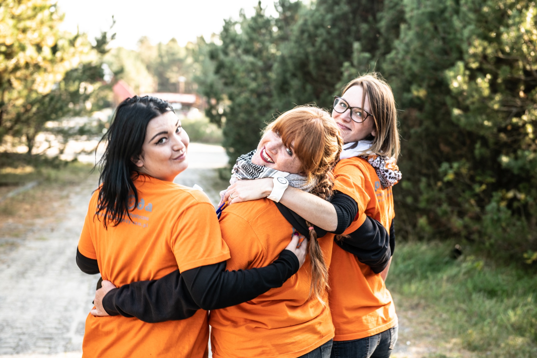 Trzy uśmiechnięte kobiety w pomarańczowych koszulkach z logo firmy, obejmujące się na tle leśnej ścieżki.