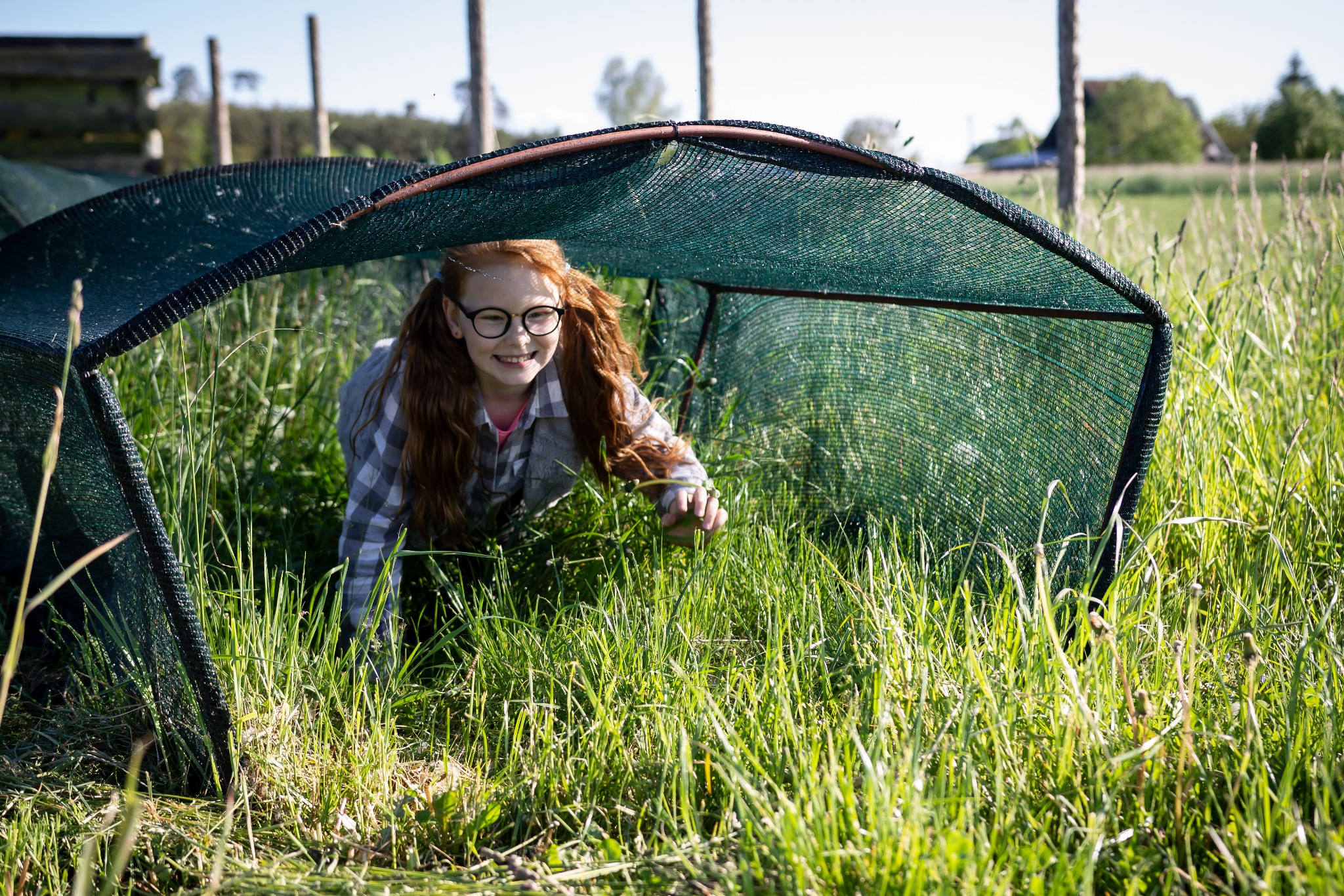 Dziewczynka z rudymi włosami i okularach uśmiecha się, wychodząc spod zielonej siatki w wysokiej trawie na łące.