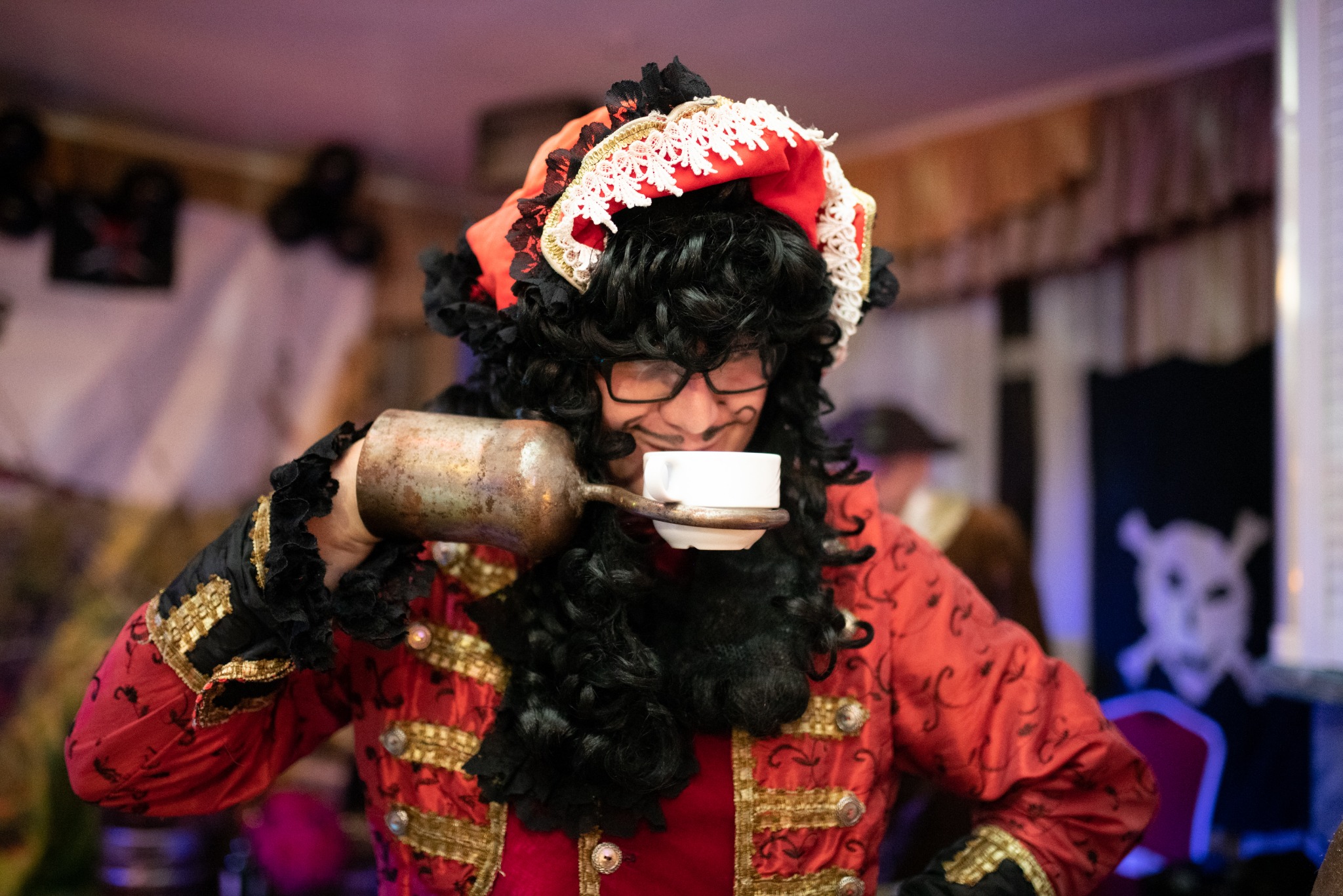 Osoba w kostiumie pirata z hakiem zamiast dłoni, nalewająca napój z metalowego dzbanka do białej filiżanki, na tle dekoracji imprezowej.