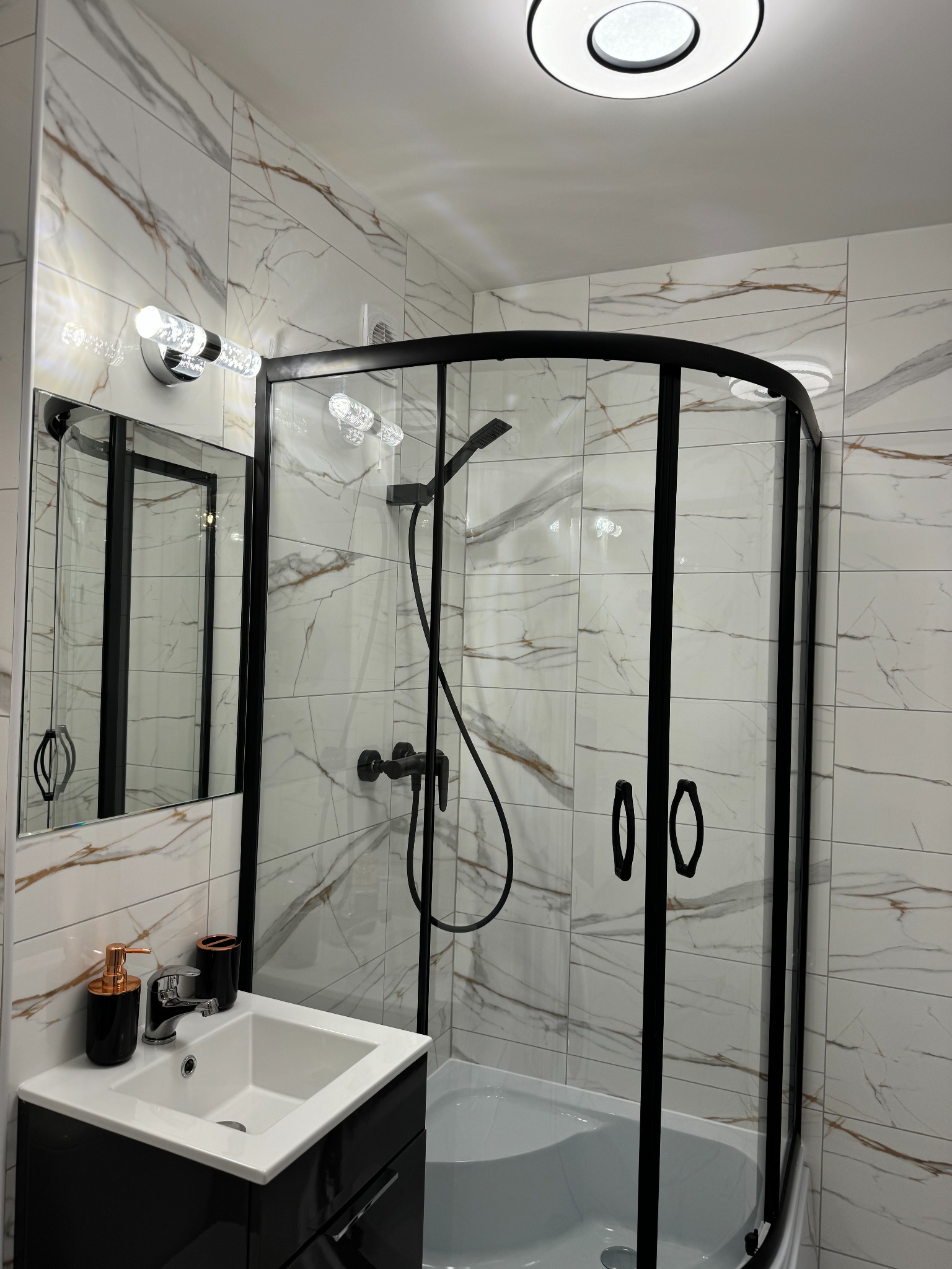 Nowoczesna łazienka z kabiną prysznicową z czarnymi profilami, umywalką nablatową na czarnej szafce i marmurowymi płytkami na ścianach.