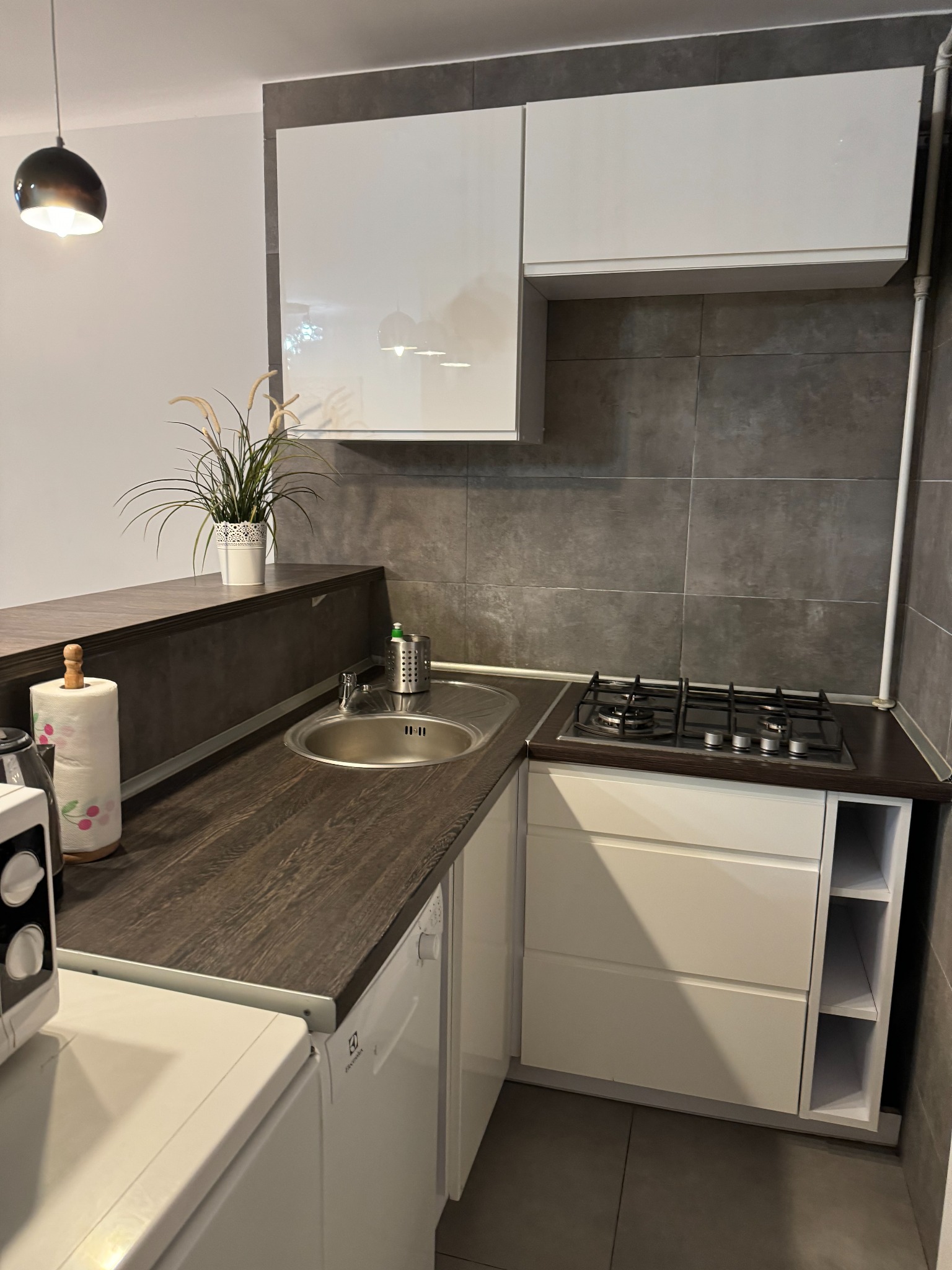 Kompaktowa kuchnia z ciemnym blatem, białymi szafkami i szarymi płytkami, oświetlona wiszącą lampą, z widocznym zlewem, kuchenką gazową i mikrofalówką Electrolux.