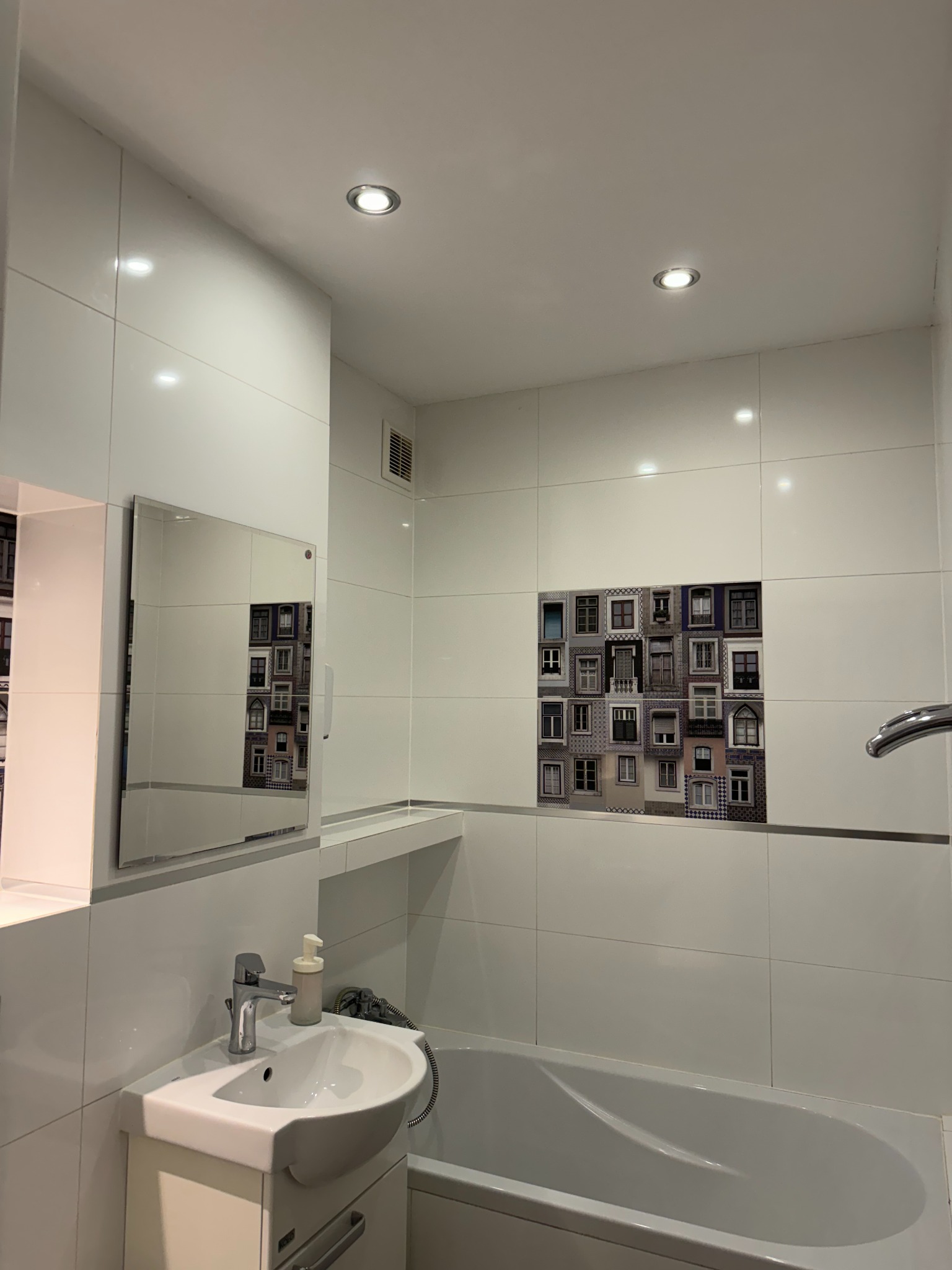 Biała łazienka z wanną, umywalką z szafką i lustrem, wykończona płytkami z motywem okien.