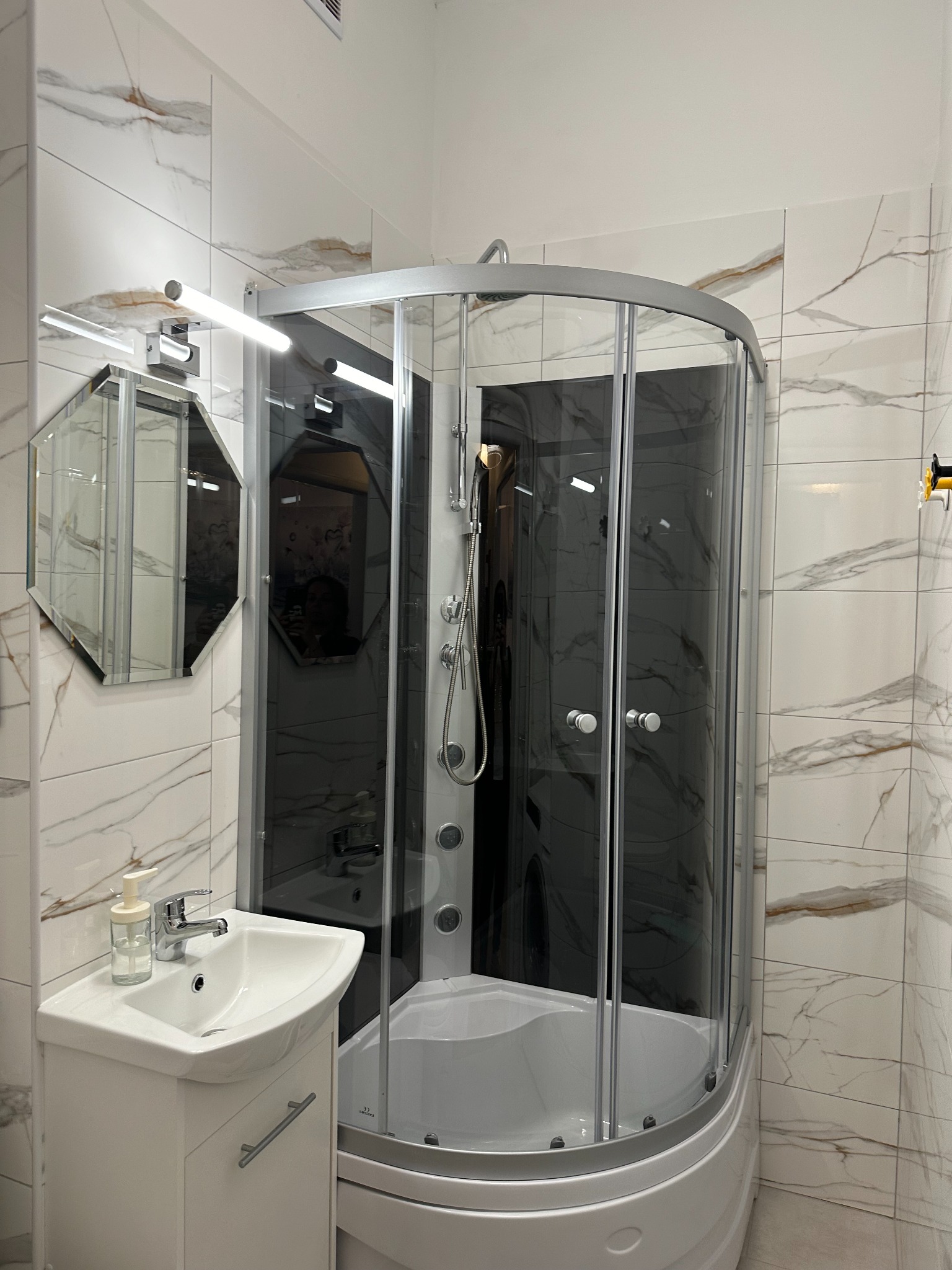 Nowoczesna łazienka z kabiną prysznicową z czarnym panelem, umywalką na szafce i lustrem o geometrycznym kształcie na marmurowych płytkach.