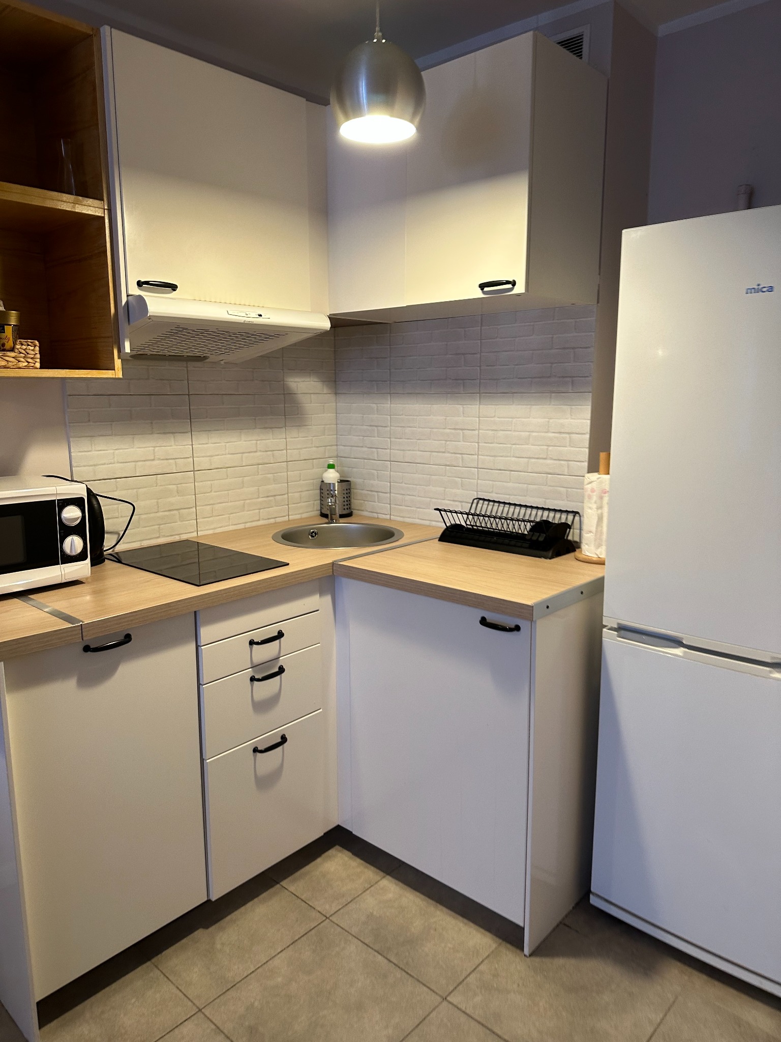 Nowoczesna, jasna kuchnia z białymi szafkami, drewnianym blatem, płytą indukcyjną, mikrofalówką i białymi cegiełkami na ścianie, oświetlona wiszącą lampą.
