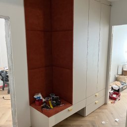 LenArt - Meble na wymiar Grzegorz Lenart - Wnęka z tapicerowanym, ceglastoczerwonym panelem ściennym zintegrowana z szafą w jasnej kolorystyce, widoczne narzędzia i akcesoria montażowe, podłoga w jodełkę.