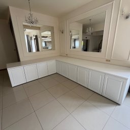 LenArt - Meble na wymiar Grzegorz Lenart - Długa, biała zabudowa meblowa z szafkami z frezowanymi frontami, dopasowana do kształtu ściany, z dwoma lustrami w ozdobnych ramach, w jasnym pomieszczeniu z błyszczącą podłogą.