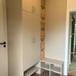 Jasna zabudowa meblowa z szafkami i wiklinowymi koszami, uzupełniona niską szafką z półkami, na tle wzorzystej podłogi i przeszklonych drzwi.