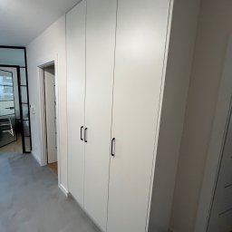 LenArt - Meble na wymiar Grzegorz Lenart - Biała szafa wnękowa z czarnymi uchwytami, widok z perspektywy korytarza, drzwi zlicowane z ścianą, szara podłoga.