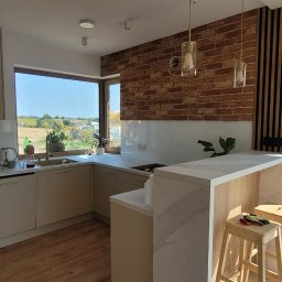 LenArt - Meble na wymiar Grzegorz Lenart - Jasna kuchnia z ceglaną ścianą i widokiem na wiejski krajobraz; drewniane stołki barowe z roboczymi rękawicami na siedzisku.