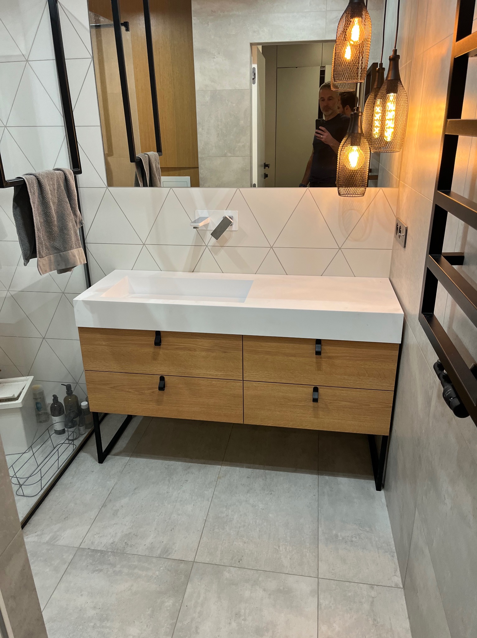 Nowoczesna łazienka w Krakowie z drewnianą szafką pod umywalkę z białym blatem, wiszącymi lampami w stylu industrialnym i geometrycznymi płytkami na ścianie.