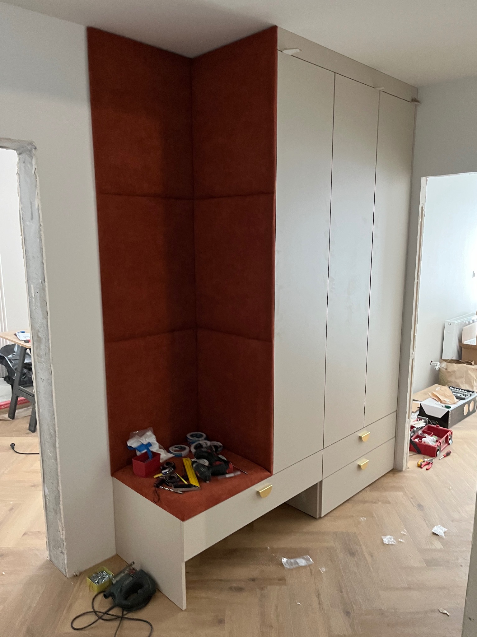 Wnęka z tapicerowanym, ceglastoczerwonym panelem ściennym zintegrowana z szafą w jasnej kolorystyce, widoczne narzędzia i akcesoria montażowe, podłoga w jodełkę.