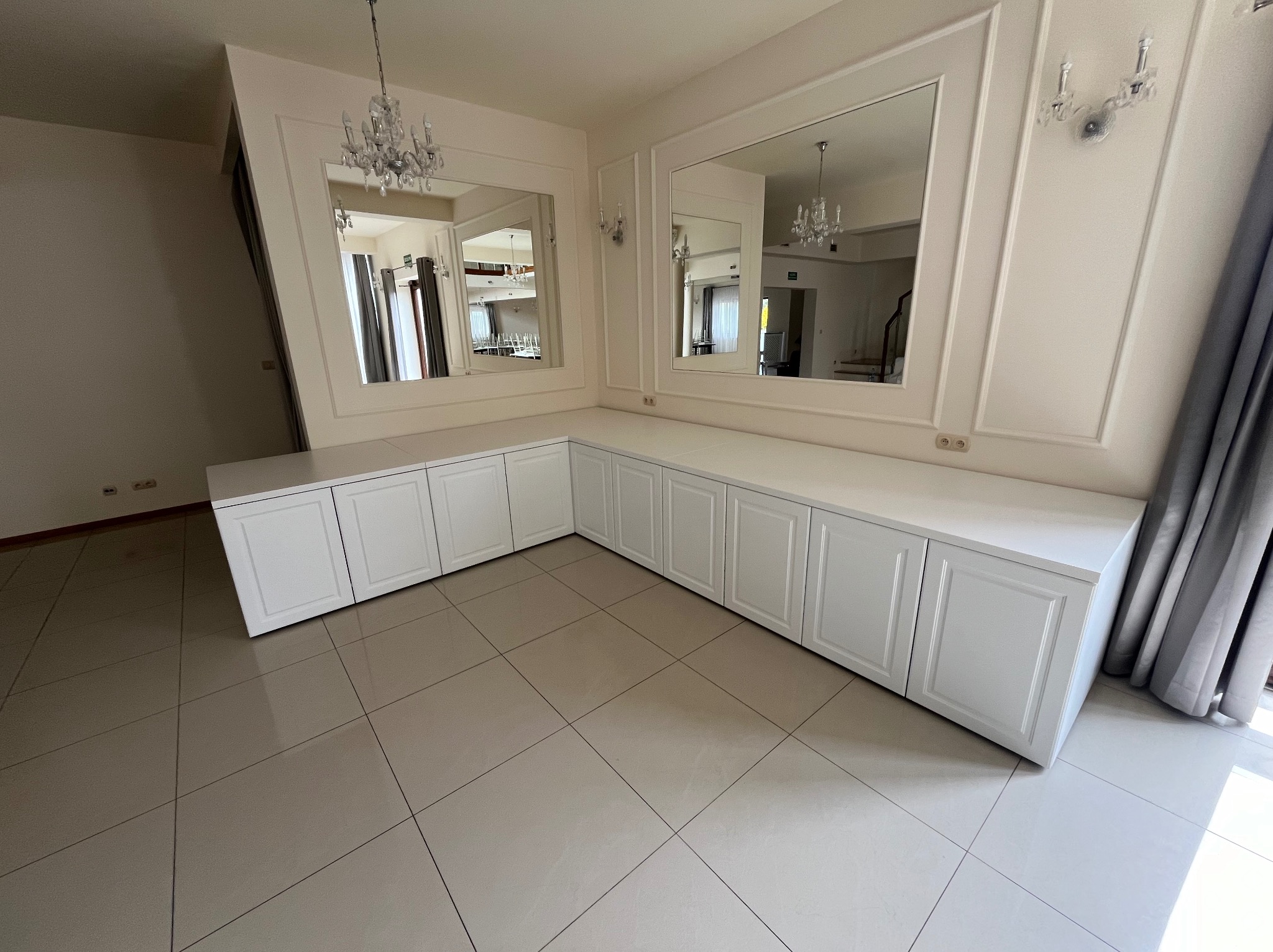 Długa, biała zabudowa meblowa z szafkami z frezowanymi frontami, dopasowana do kształtu ściany, z dwoma lustrami w ozdobnych ramach, w jasnym pomieszczeniu z błyszczącą podłogą.