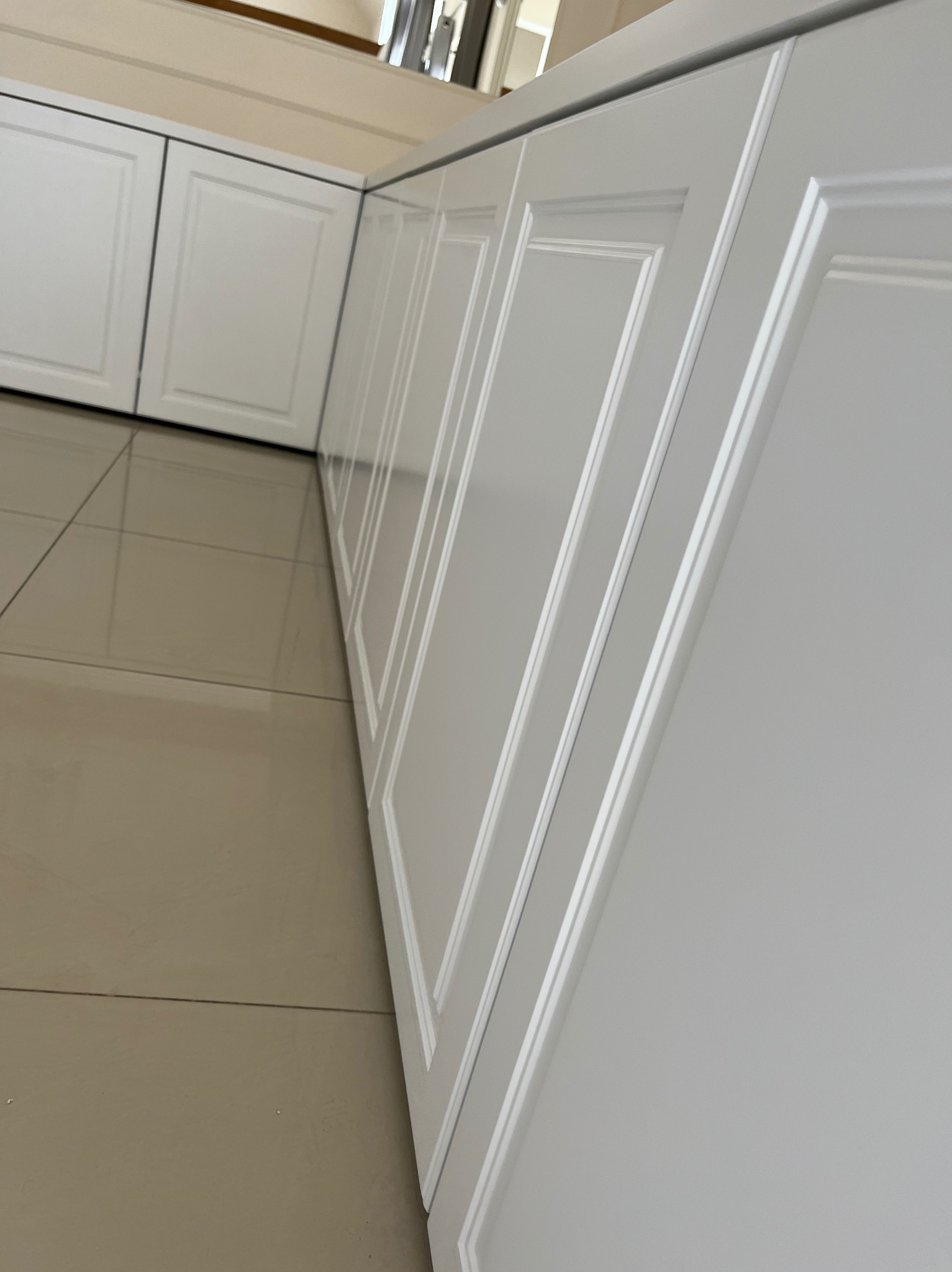 Białe fronty szafek kuchennych z frezowanymi ramkami, widok z dołu, na pierwszym planie płytki podłogowe w odcieniach beżu.