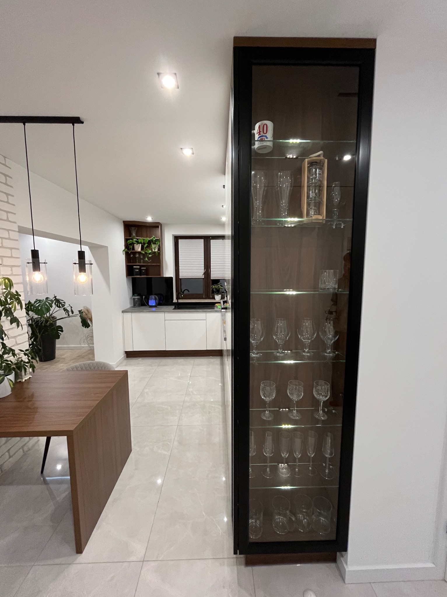 Nowoczesna kuchnia z białymi szafkami, drewnianym blatem i przeszkloną witryną z kolekcją kieliszków, widok z jadalni z drewnianym stołem.