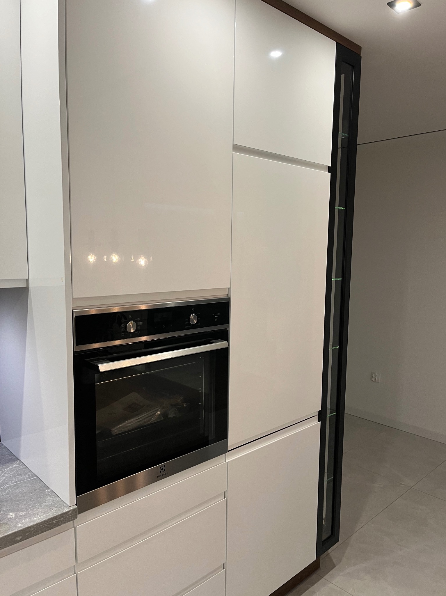 Białe, lakierowane fronty zabudowy kuchennej z piekarnikiem marki Electrolux. Widoczna szklana witryna z ciemną ramą i drewnianym akcentem u góry. Szare płytki podłogowe i fragment blatu.