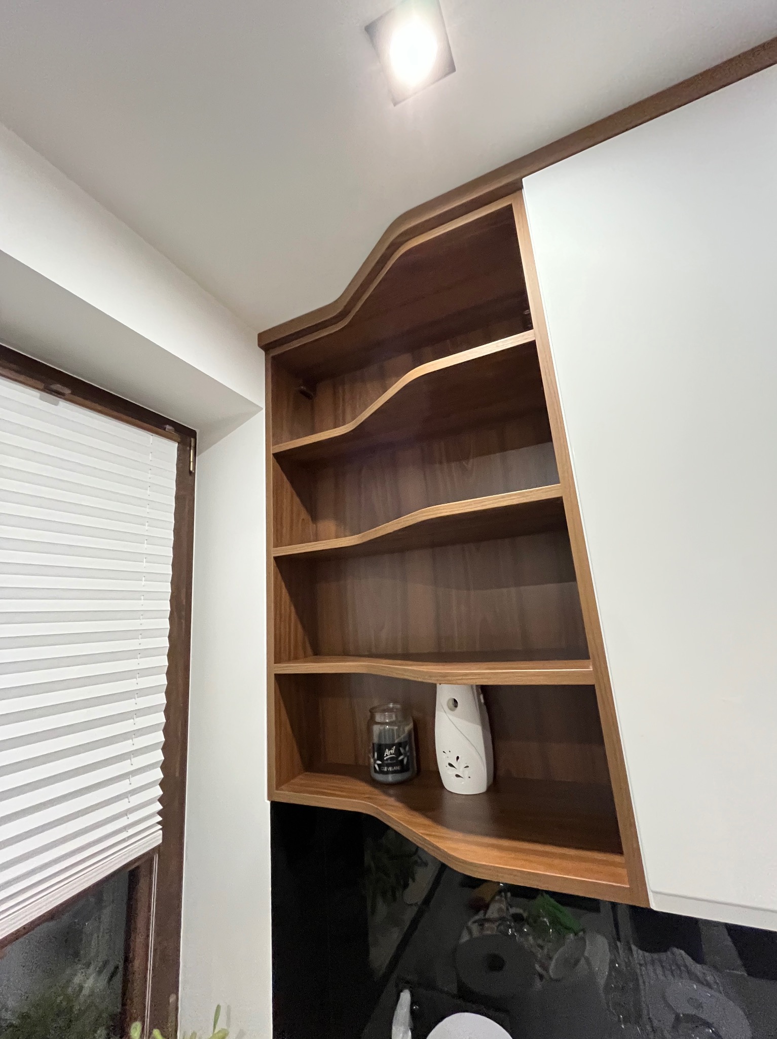 Półki z ciemnego drewna o falistych krawędziach, wkomponowane w białą zabudowę kuchenną, obok okna z żaluzjami.