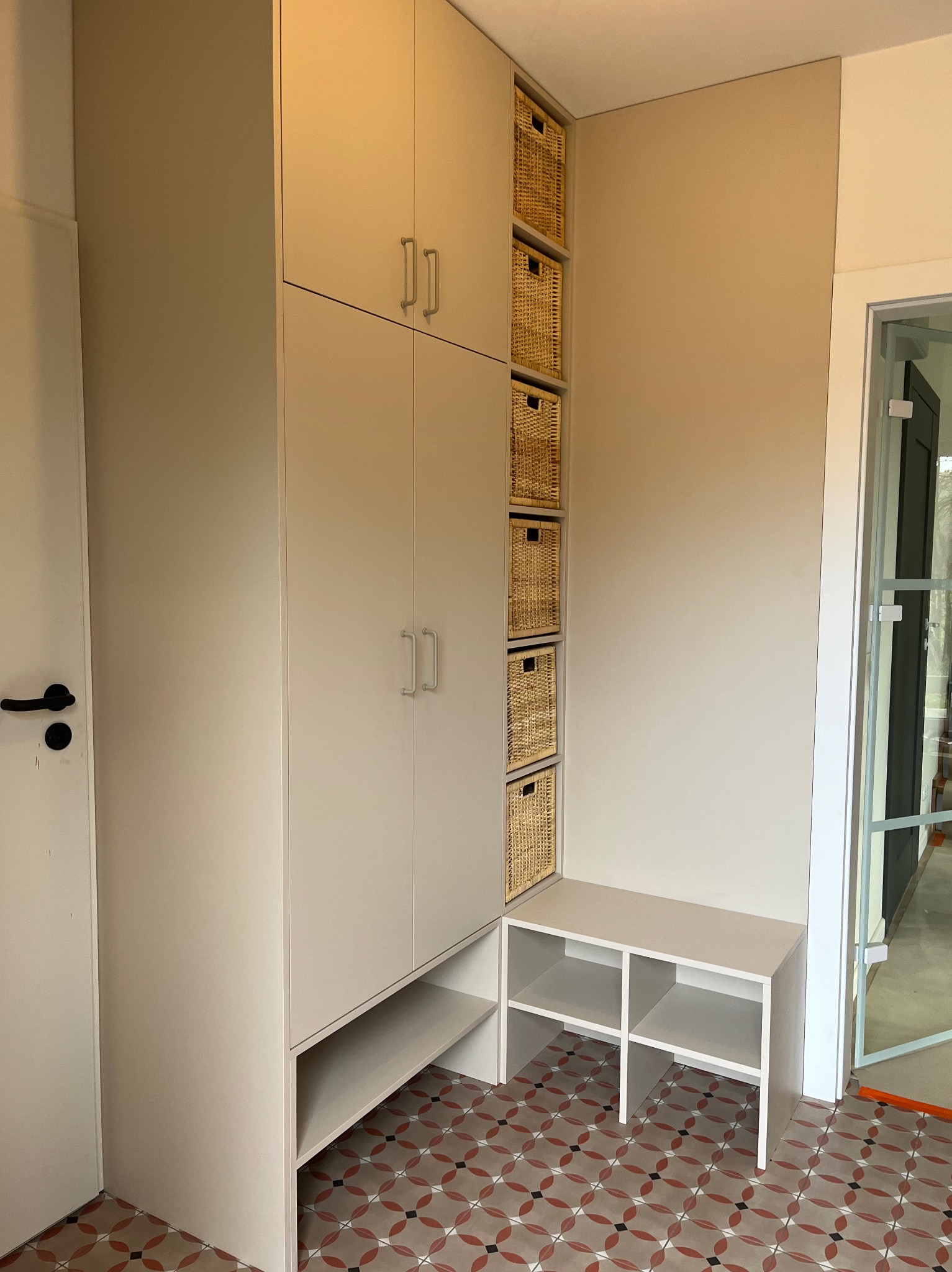 Jasna zabudowa meblowa z szafkami i wiklinowymi koszami, uzupełniona niską szafką z półkami, na tle wzorzystej podłogi i przeszklonych drzwi.