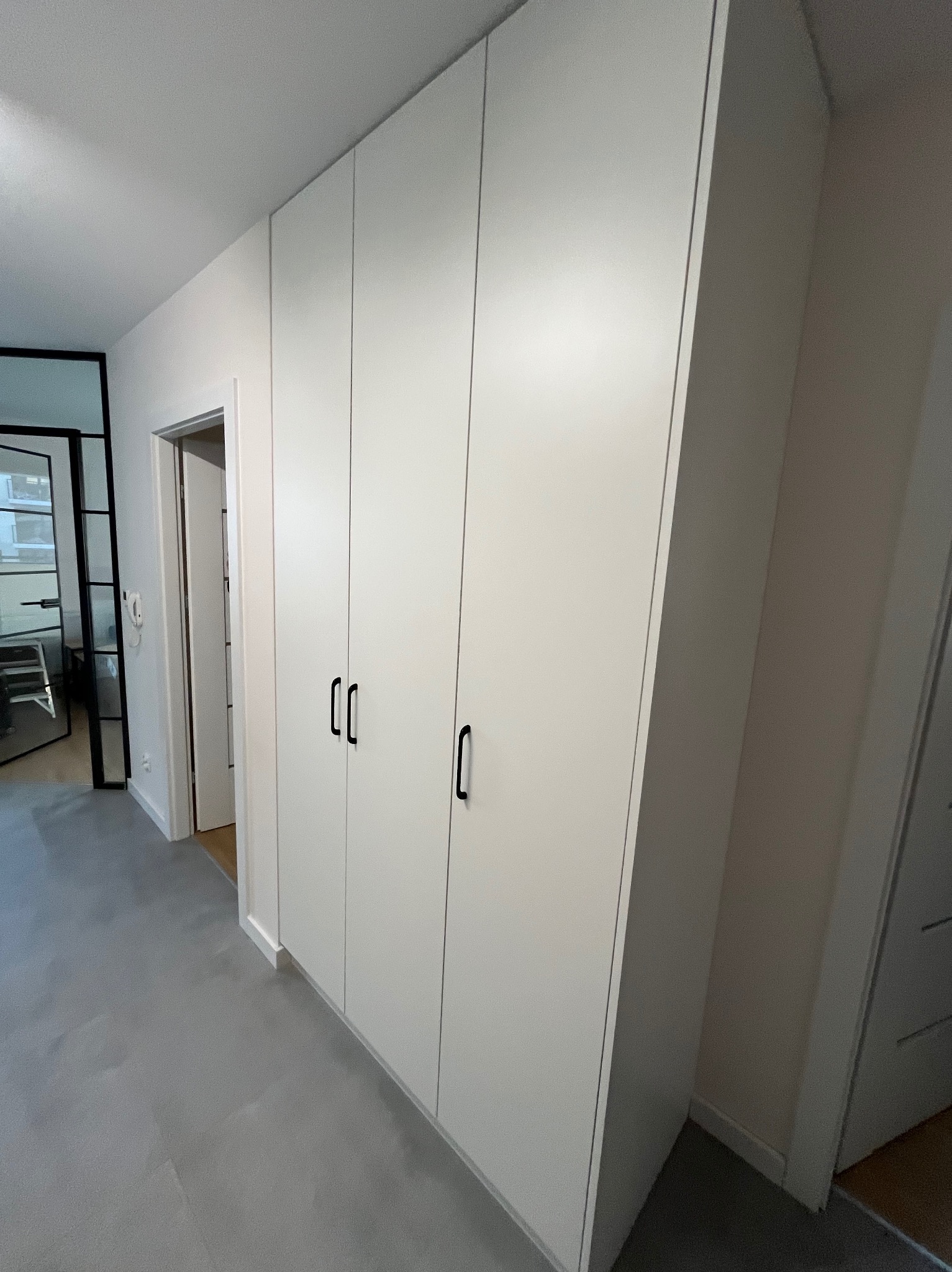 Biała szafa wnękowa z czarnymi uchwytami, widok z perspektywy korytarza, drzwi zlicowane z ścianą, szara podłoga.