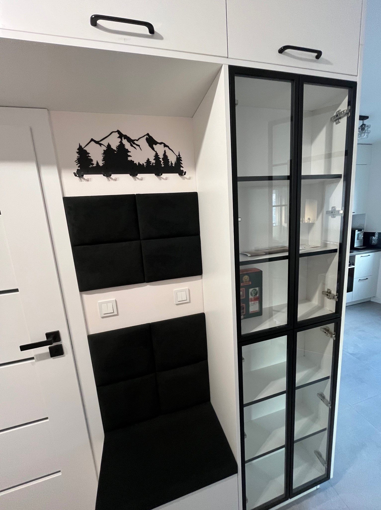 Biała zabudowa meblowa z czarnymi akcentami: przeszklona witryna z czarnymi szprosami, siedzisko z czarnych paneli tapicerowanych, dekoracja ścienna z motywem górskim.