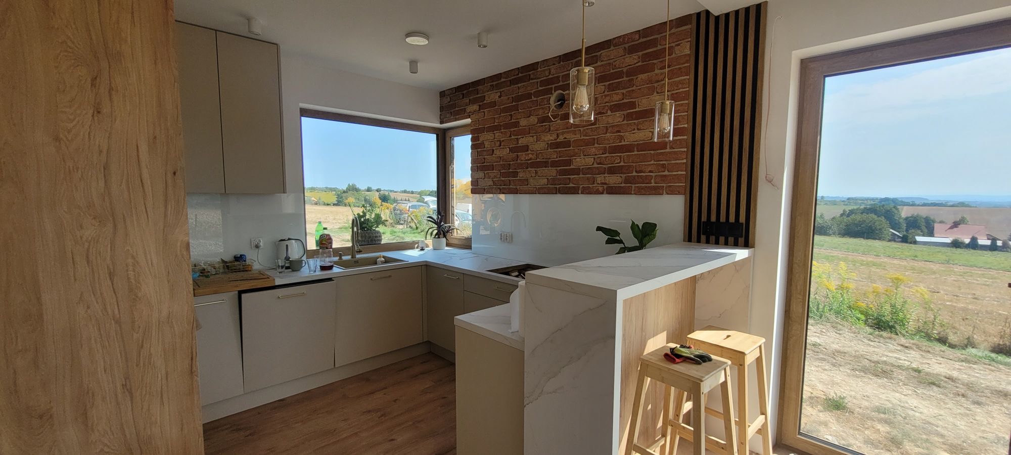 Jasna kuchnia z ceglaną ścianą i widokiem na wiejski krajobraz; drewniane stołki barowe z roboczymi rękawicami na siedzisku.