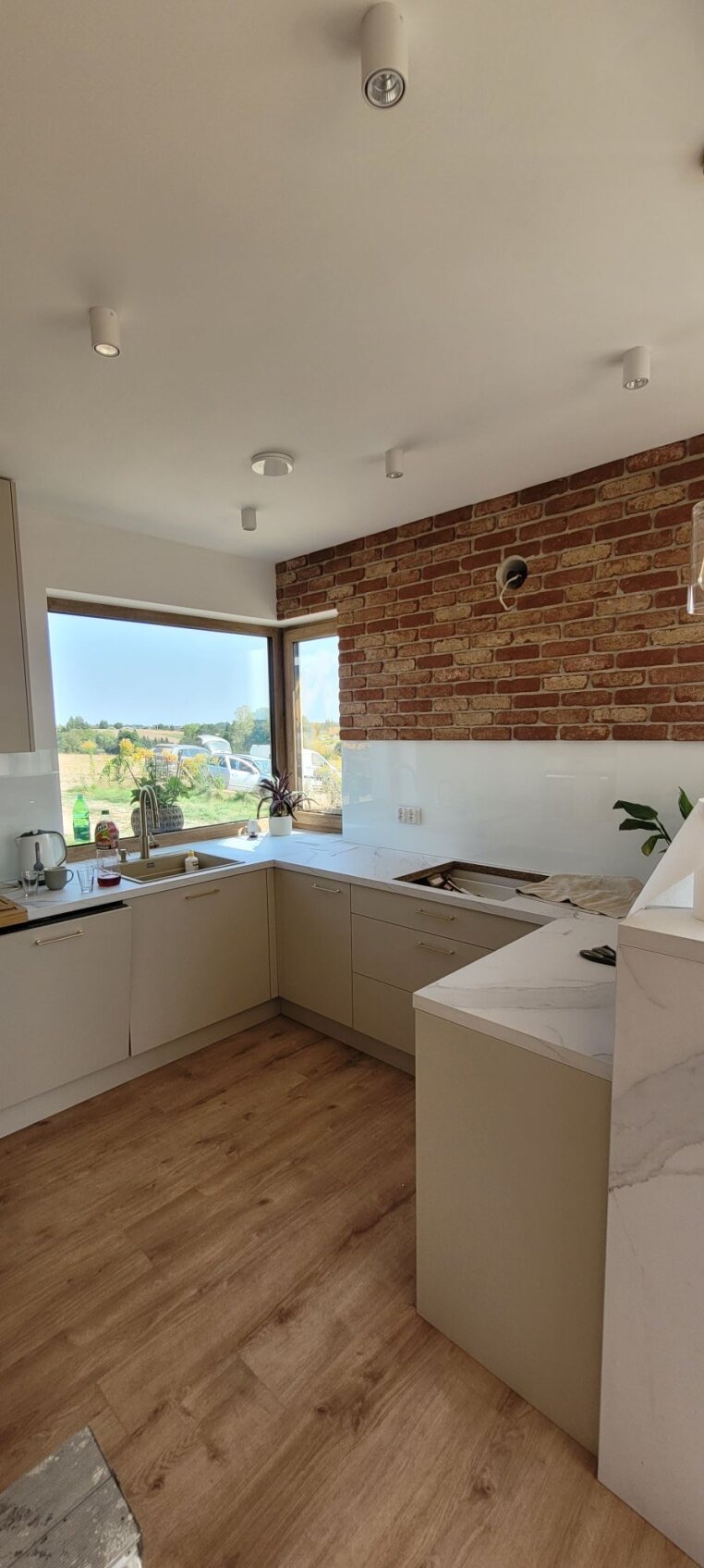 Jasna kuchnia z drewnianą podłogą i ceglaną ścianą, widok na zewnątrz przez duże okno, brakujące elementy blatu, w tle krajobraz.