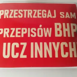 Czerwona tablica informacyjna z napisem 'PRZESTRZEGAJ SAM PRZEPISÓW BHP I UCZ INNYCH!' w kolorze kremowym, przymocowana dwoma nitami.
